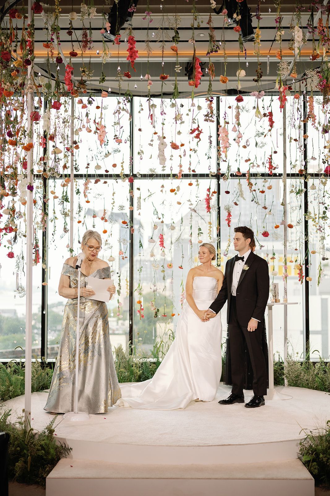 Wedding couple on an outdoor stage beneath hanging floral decorations, with a violinist in a silver dress nearby