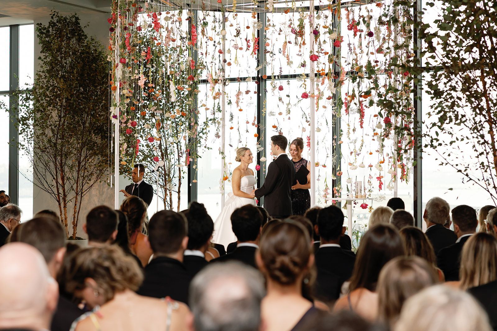 Wedding ceremony with couple and officiant under hanging floral greenery, guests seated in front.