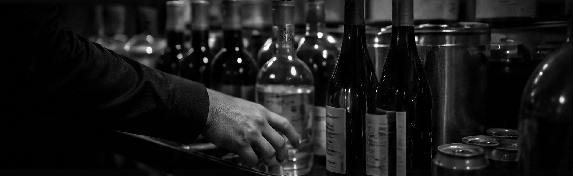 A hand reaches for glasses among liquor bottles on a dark bar shelf.