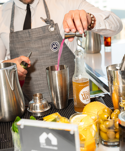 Bartender pouring pink drink into metal shaker with lemons and bottles on a bar counter