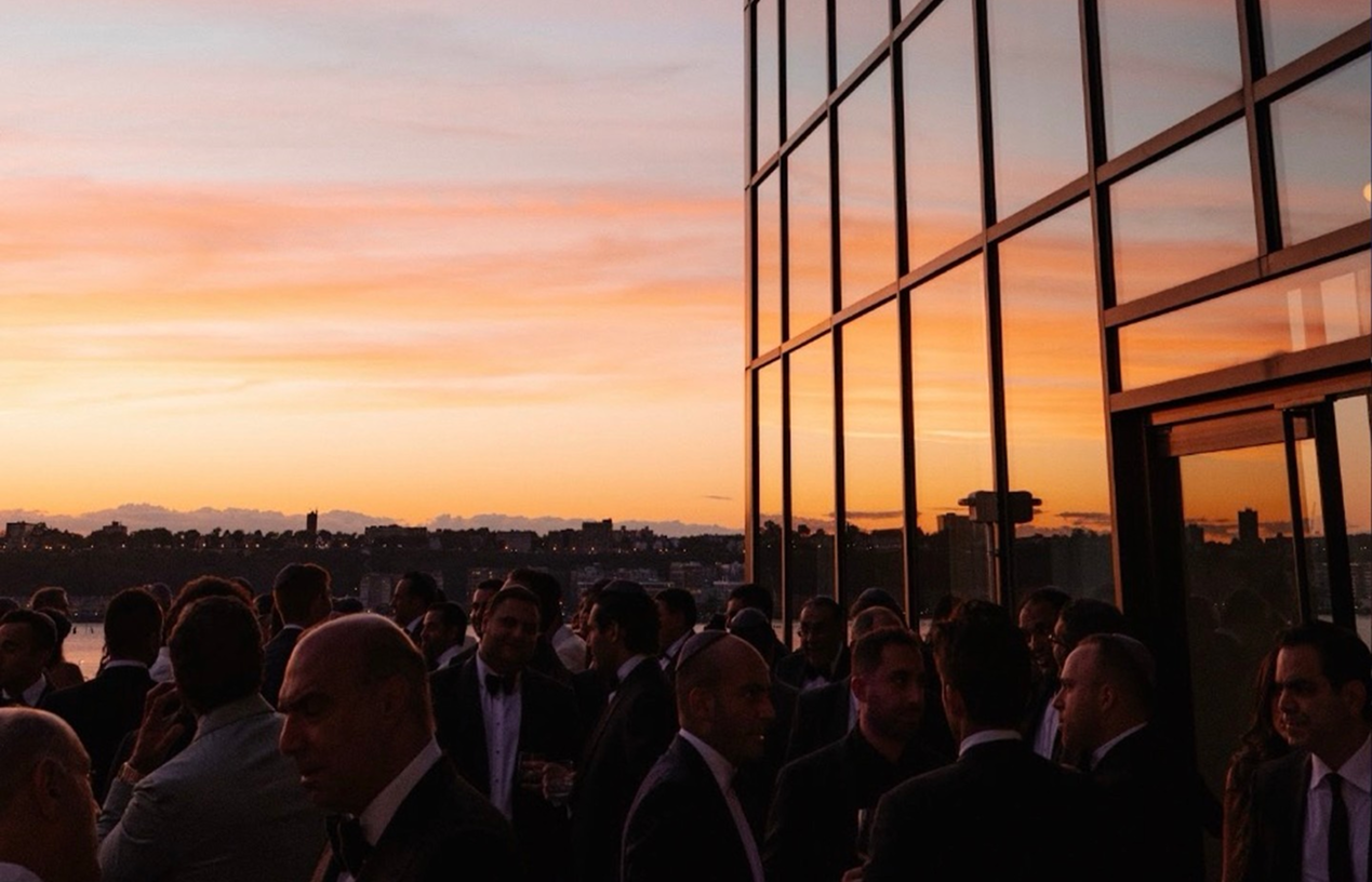 Crowd gathered on a terrace at sunset beside a glass-walled building.
