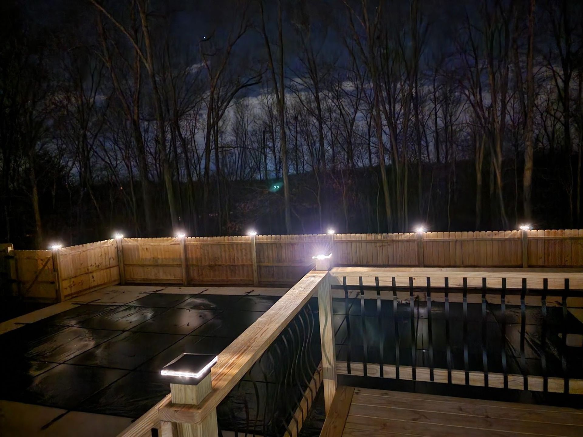 A backyard pool deck at night, illuminated by solar lights along the fence and railing, with trees in the dark background.