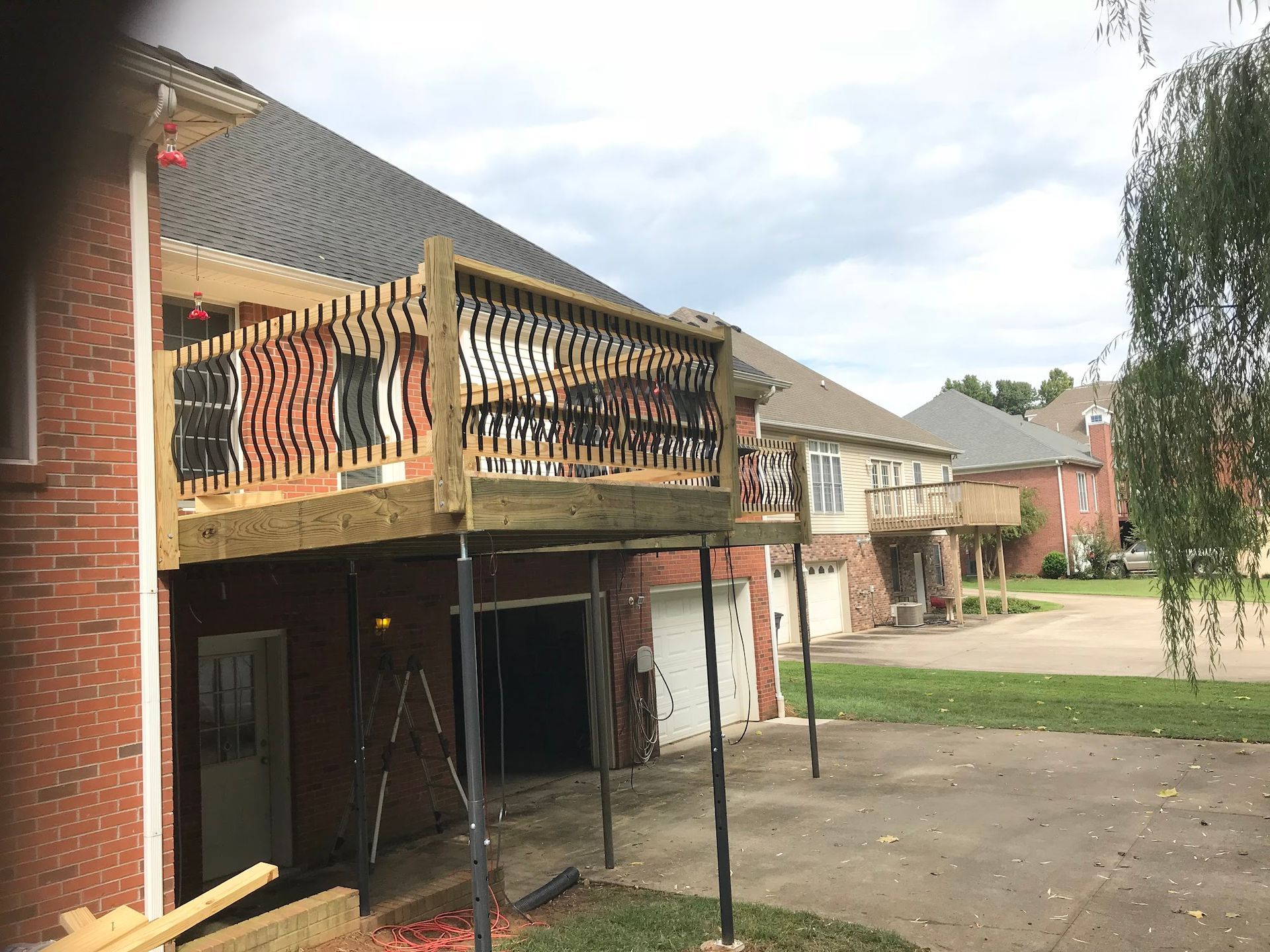 An elevated wooden deck under construction attached to the back of a brick house with a concrete driveway below.