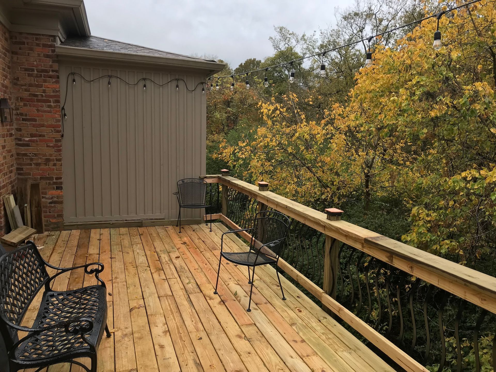 A newly built wooden deck with a black bench and chair, attached to a brick house overlooking a wooded area in autumn.