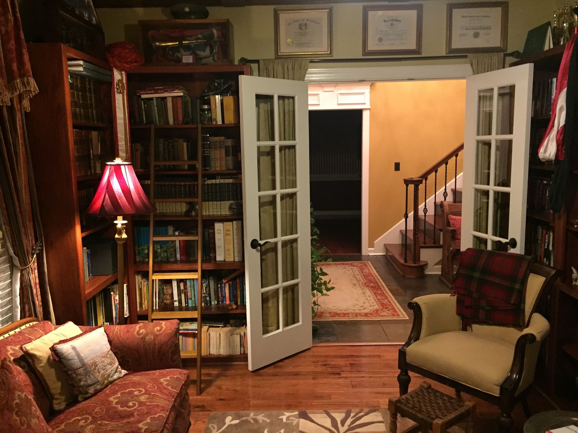 A cozy home library with floor-to-ceiling bookshelves, a reading chair, a red lamp, and French doors leading to a hallway.