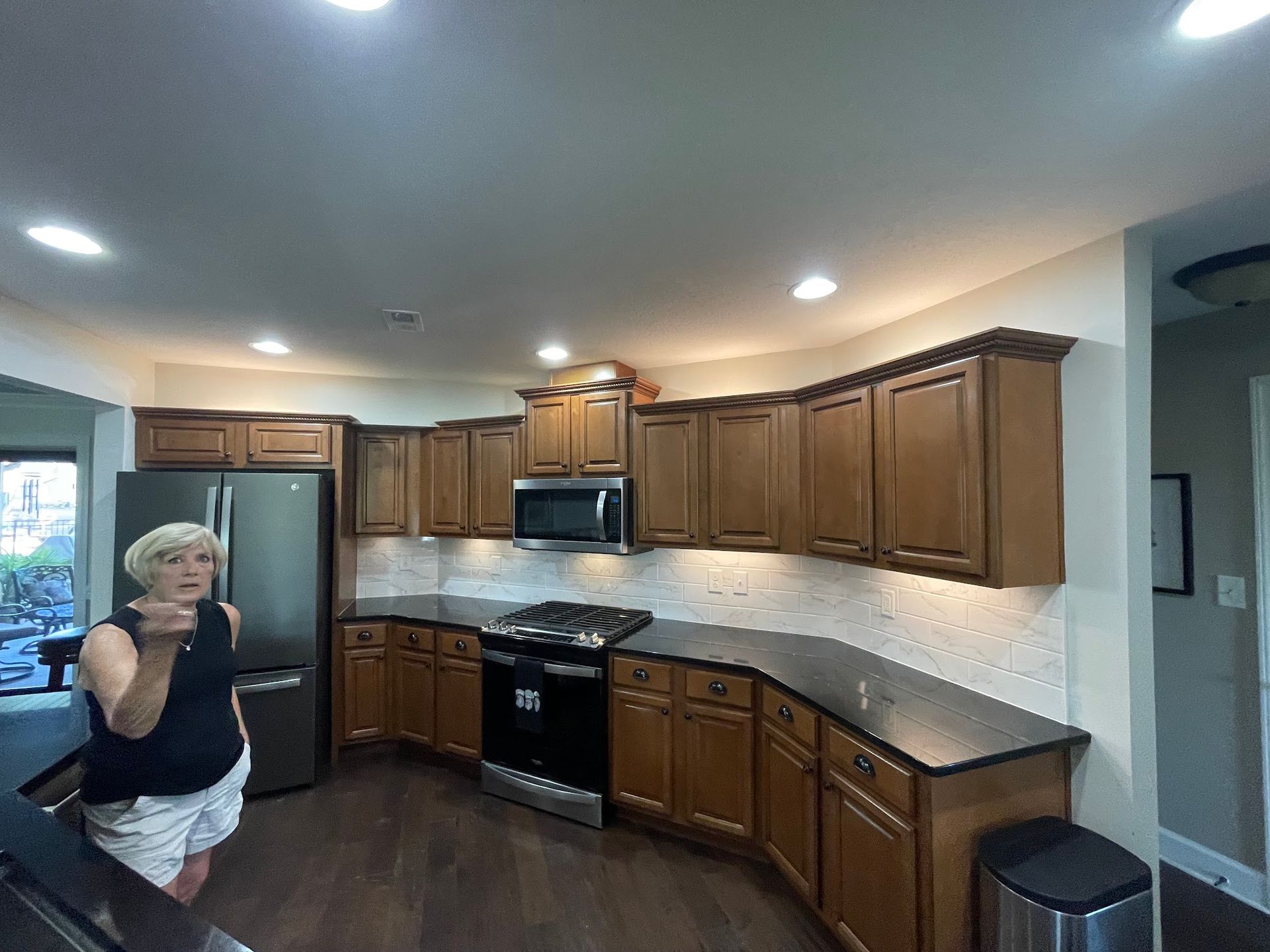 A person stands in a kitchen featuring medium-tone wood cabinets, black countertops, stainless appliances, and tile.