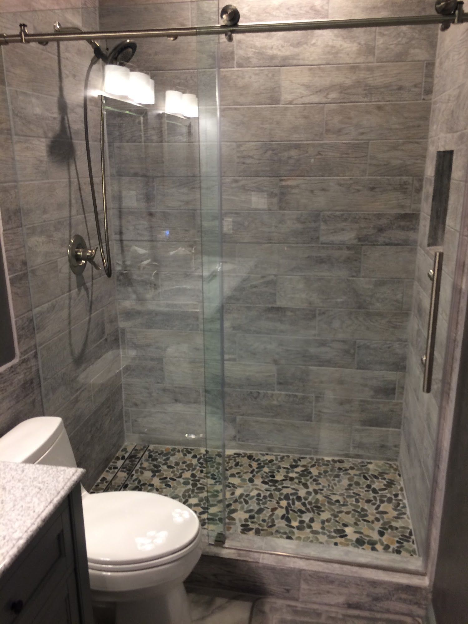 A glass-enclosed shower with gray rectangular wall tiles and a pebble-stone floor next to a white toilet.