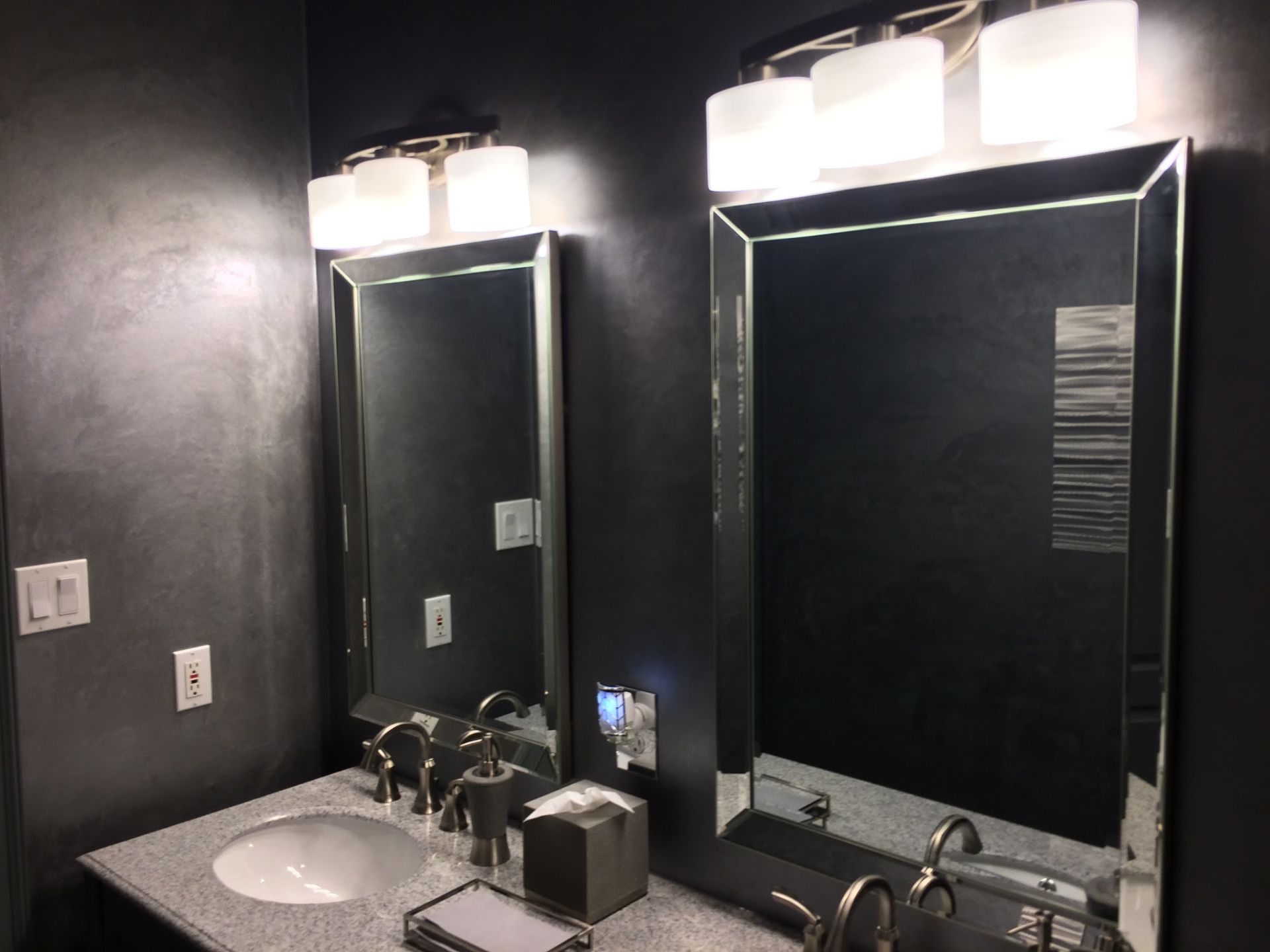 A dual-sink bathroom vanity with dark walls, matching framed mirrors, and modern glass-shaded light fixtures.