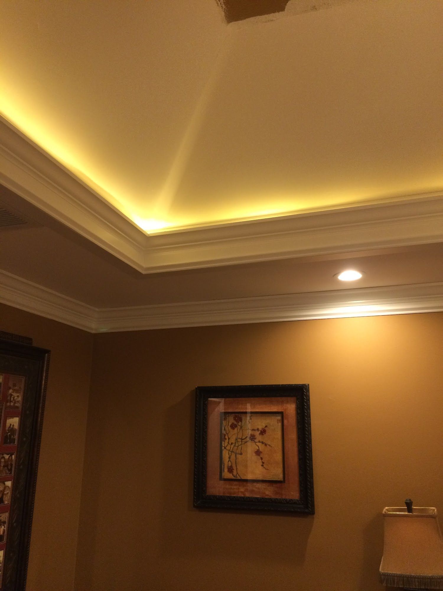An interior view of a room with golden wall paint, crown molding with accent lighting, and a framed wall art piece.
