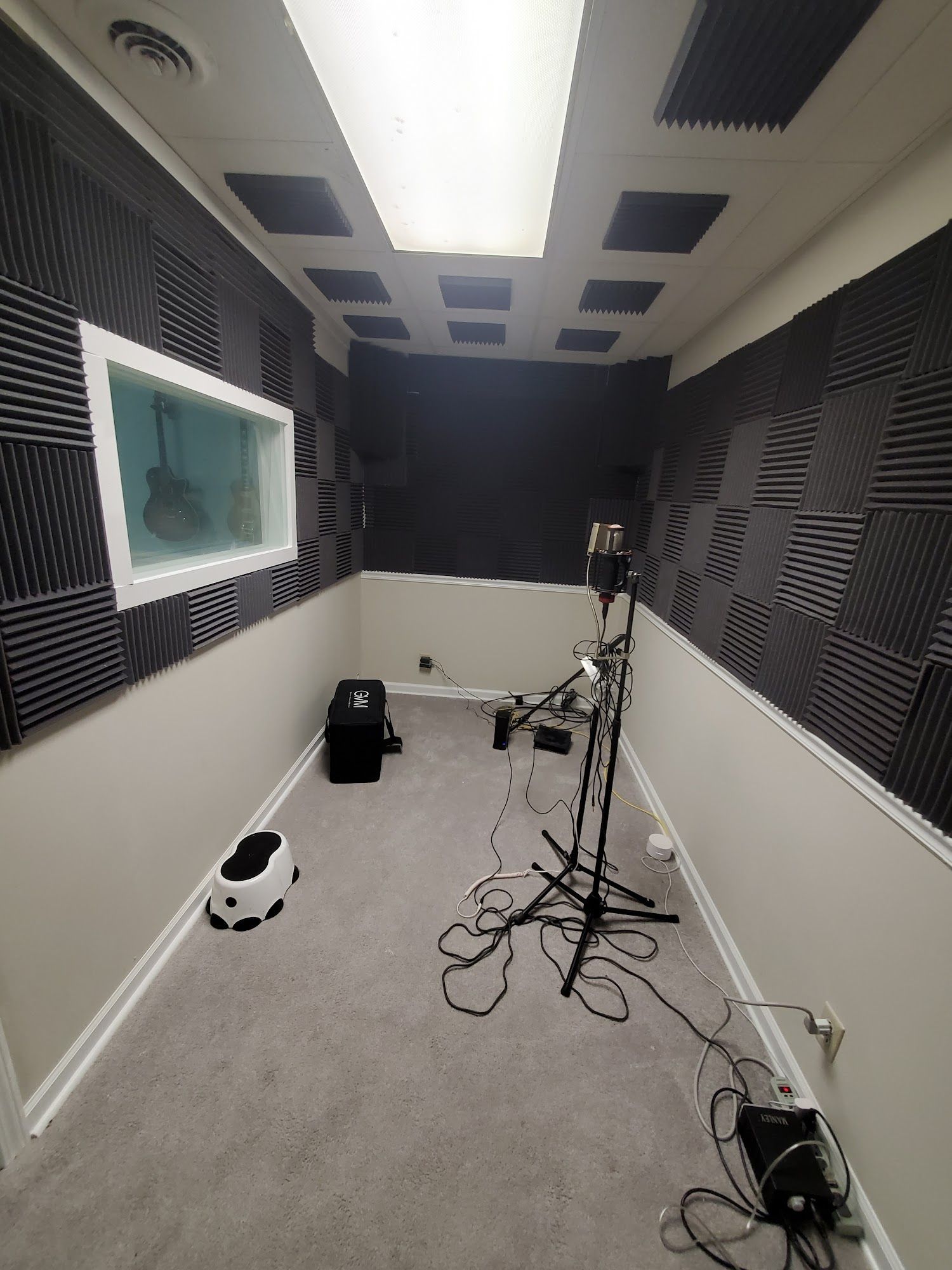 A small recording booth featuring acoustic foam panels on walls and ceiling, a microphone stand, and audio equipment.