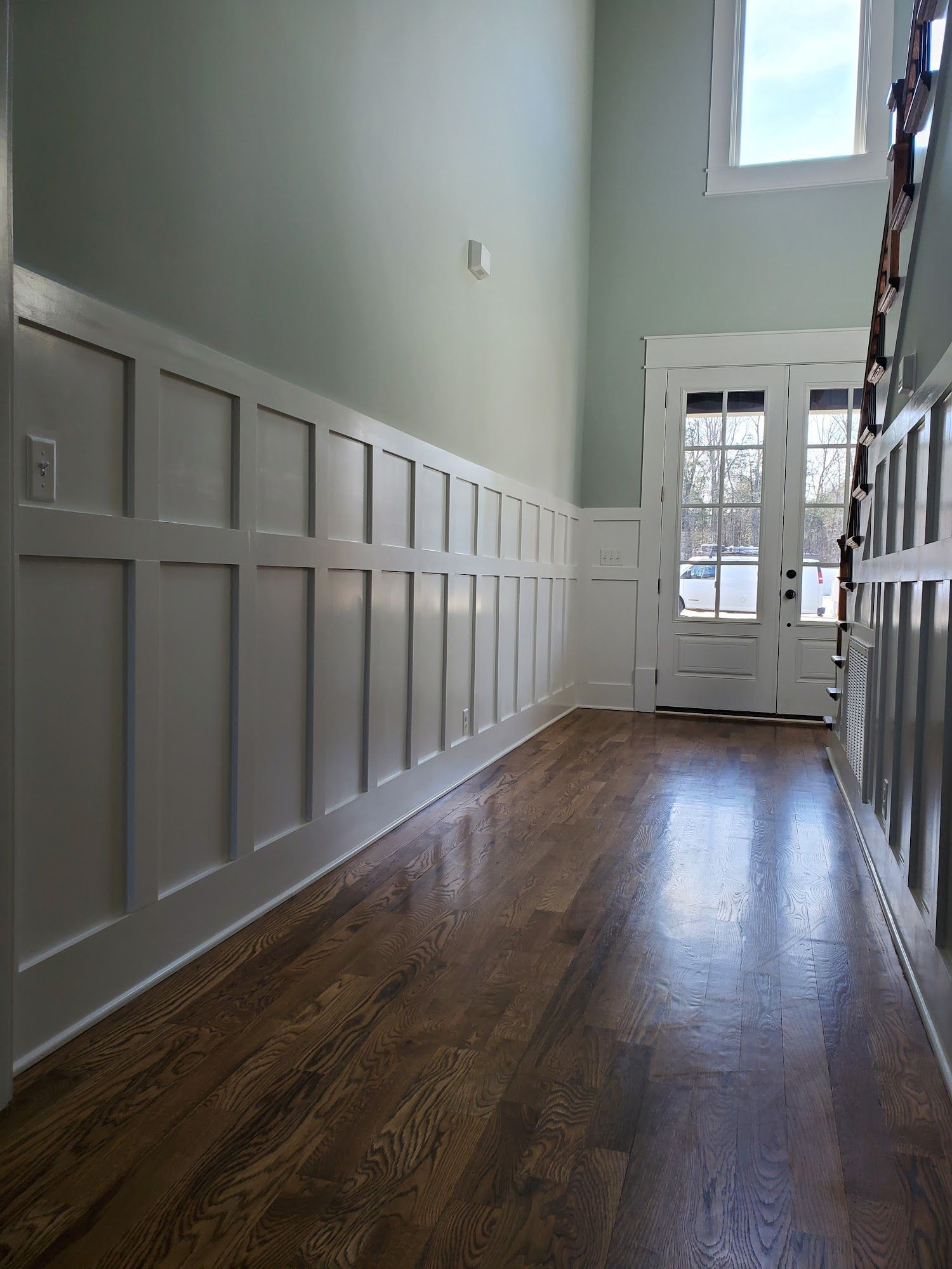 A hallway with light green walls, white wainscoting, and dark hardwood floors leading toward a glass-paneled front door.