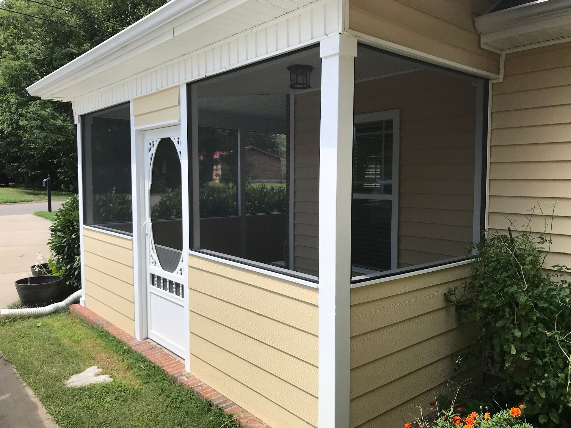 A screened-in porch with a white door and pale yellow siding, attached to a house next to a small garden.