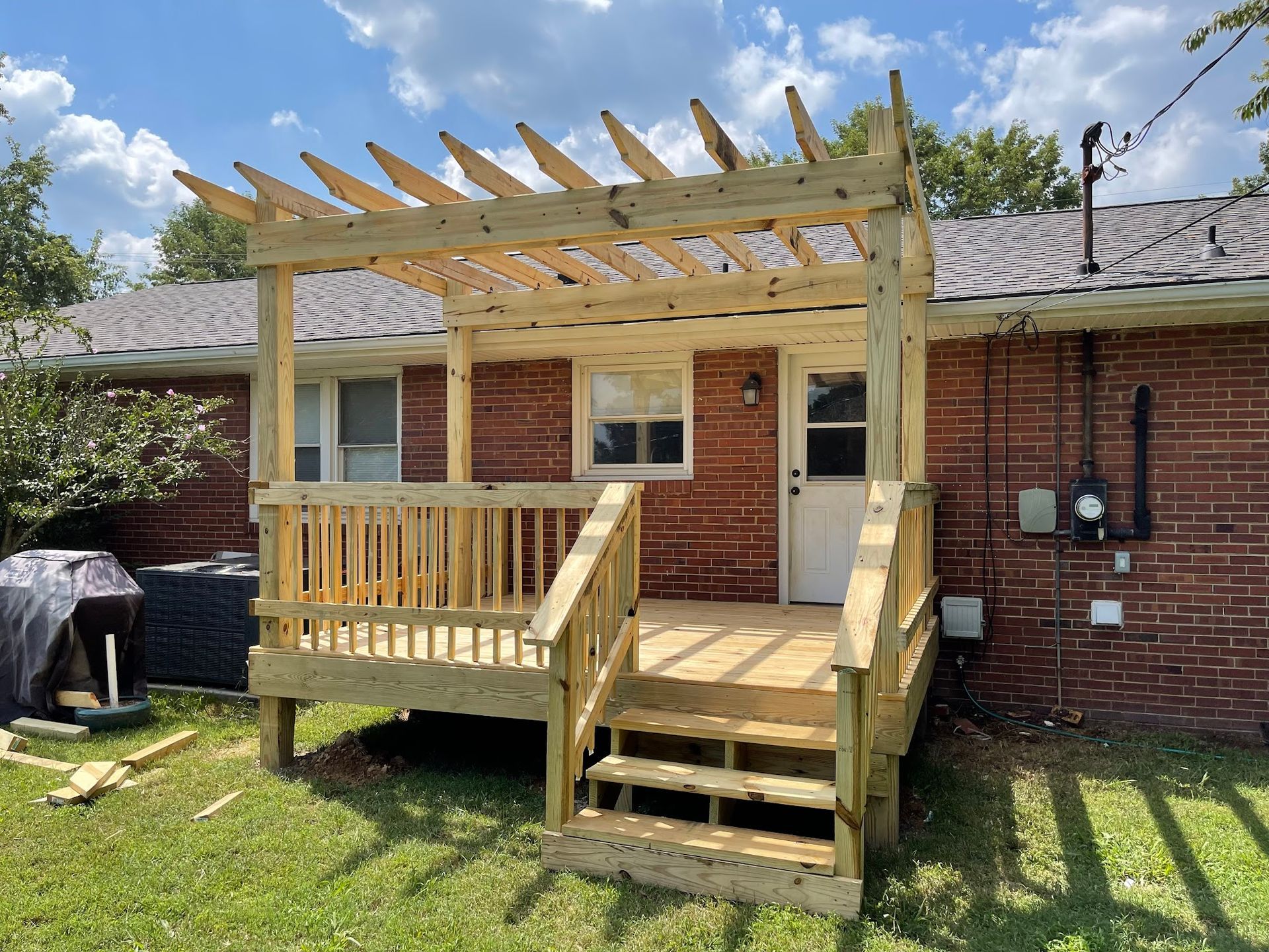 A newly constructed wooden deck and pergola attached to the rear of a brick house with steps leading down to the lawn.