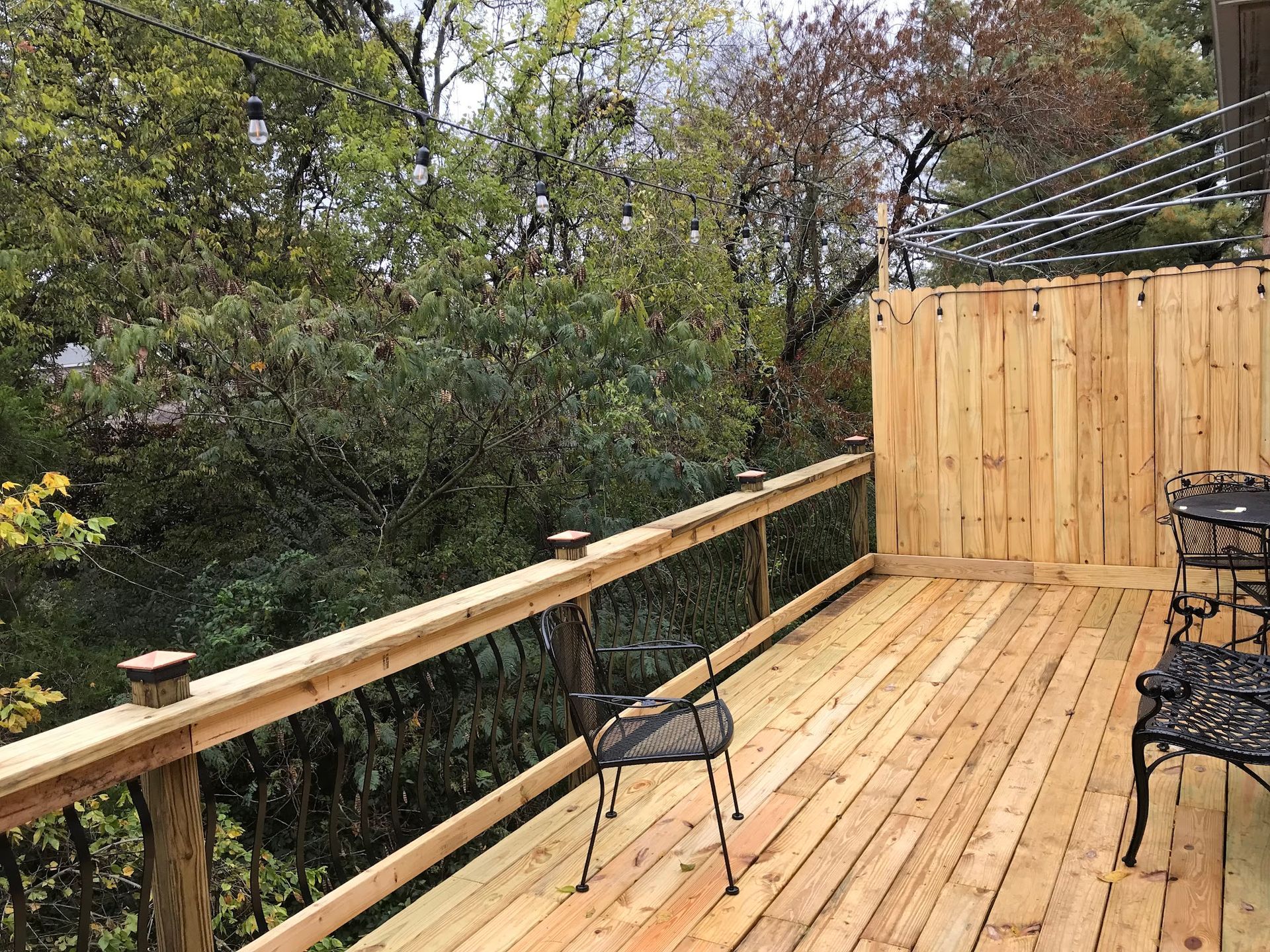 A wooden deck with a railing and privacy wall, overlooking a dense, leafy forest with a few chairs arranged on the planks.