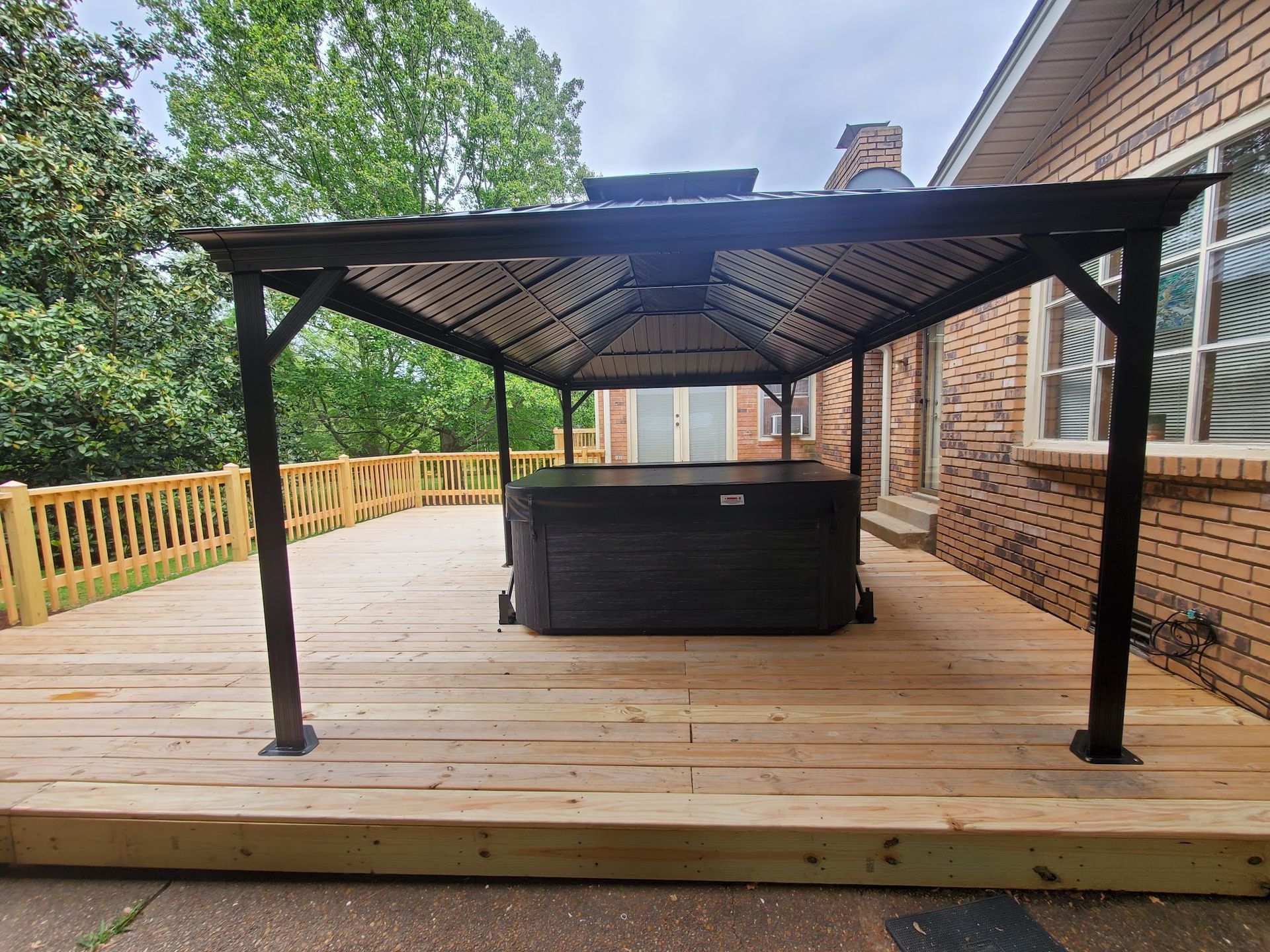 A black hot tub covered by a dark metal gazebo on a newly built wooden deck next to a brick house.