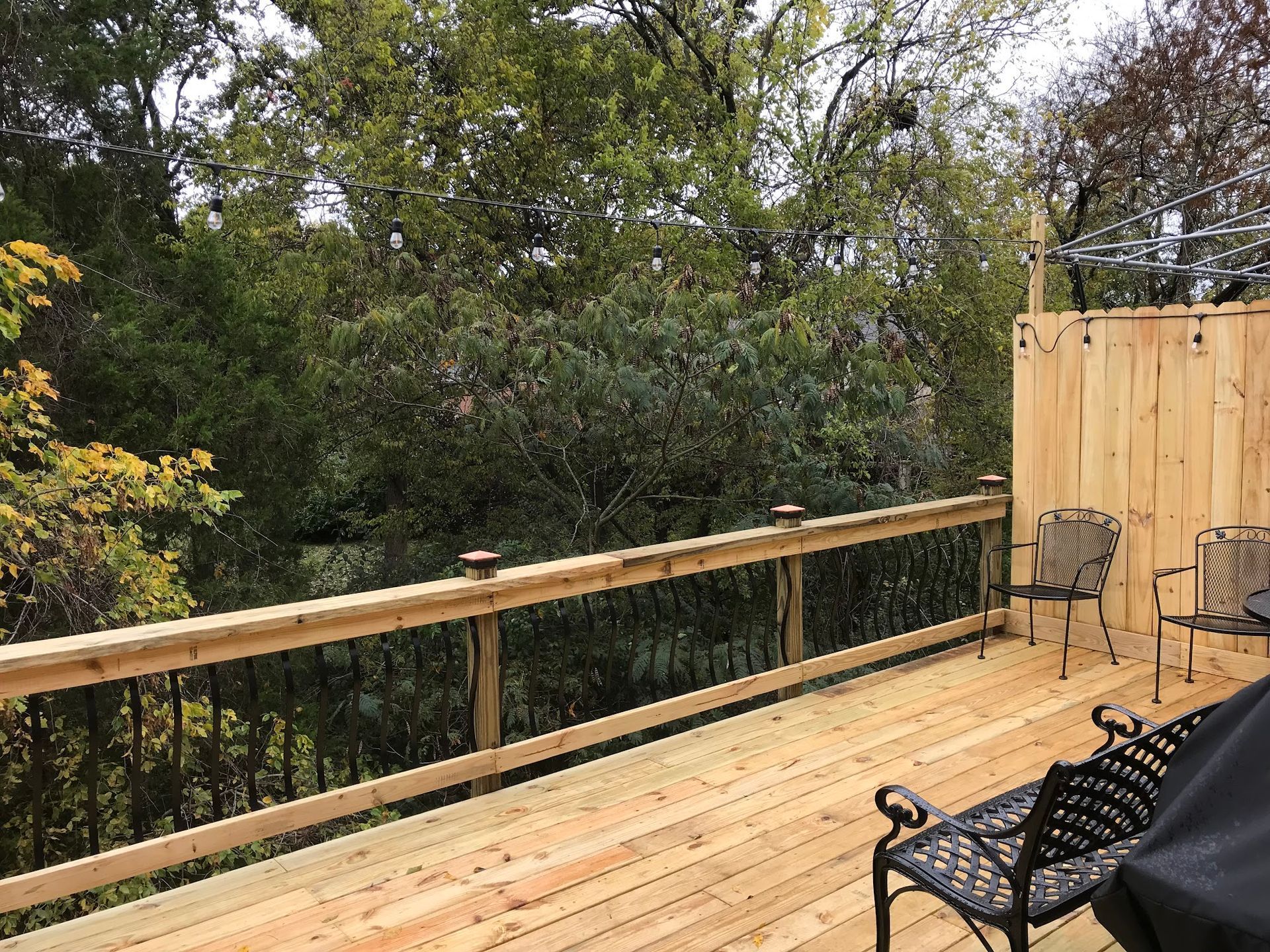 A wooden deck with a railing and privacy screen overlooking a backdrop of dense green trees.