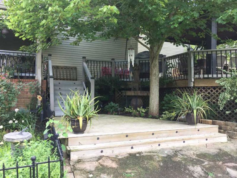 A wooden deck with a staircase leads up to a covered porch area with potted plants under a large tree in a backyard.