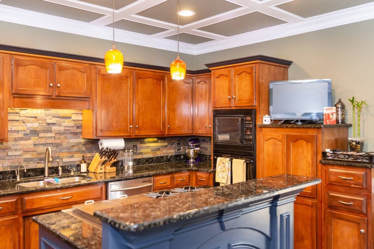 A kitchen with wooden cabinets, stone backsplash, dark granite countertops, and a blue island under a coffered ceiling.