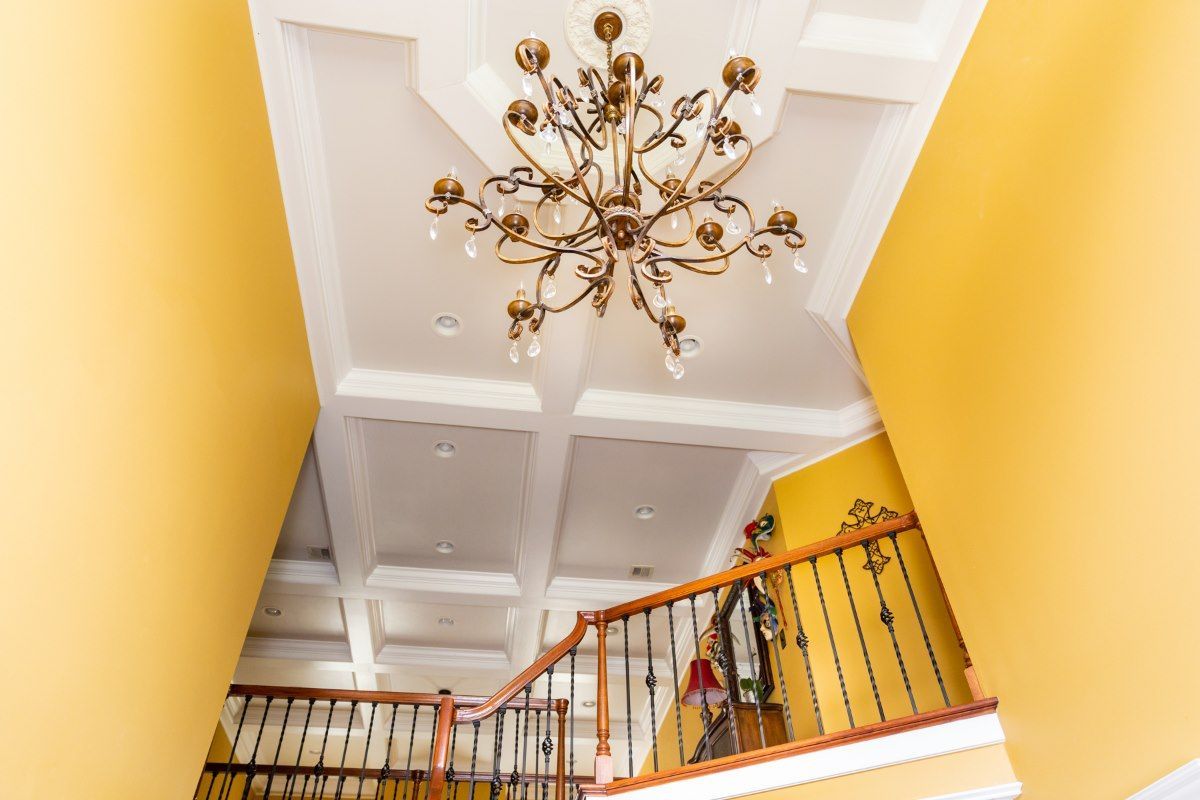 A grand foyer with yellow walls, a white coffered ceiling, and an ornate gold chandelier above a wooden staircase.