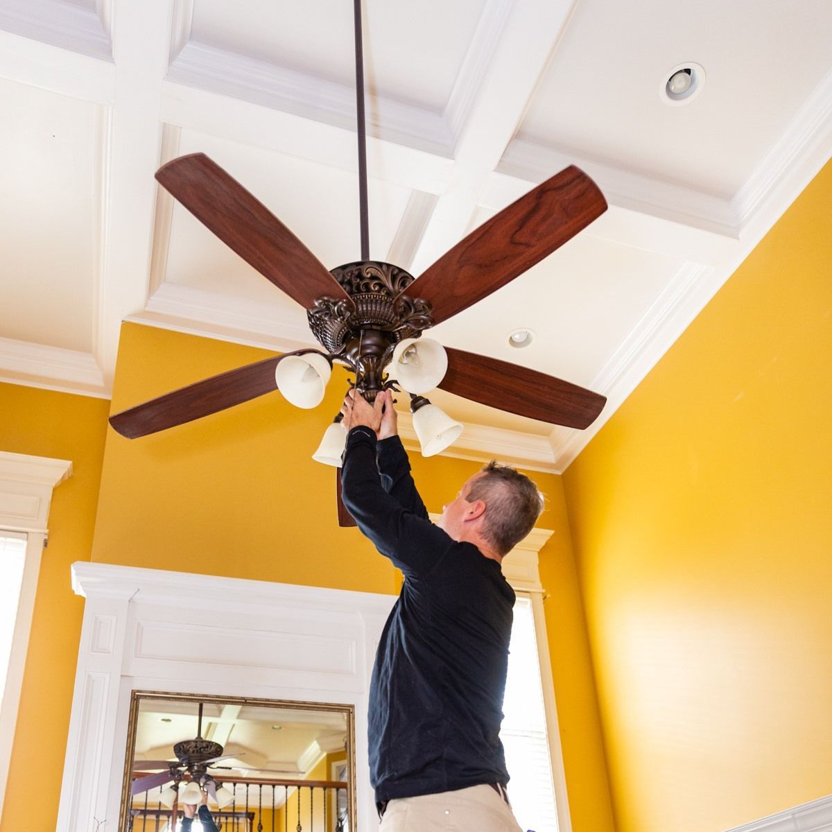 A person standing on a ladder adjusts a wooden-bladed ceiling fan in a room with bright yellow walls.
