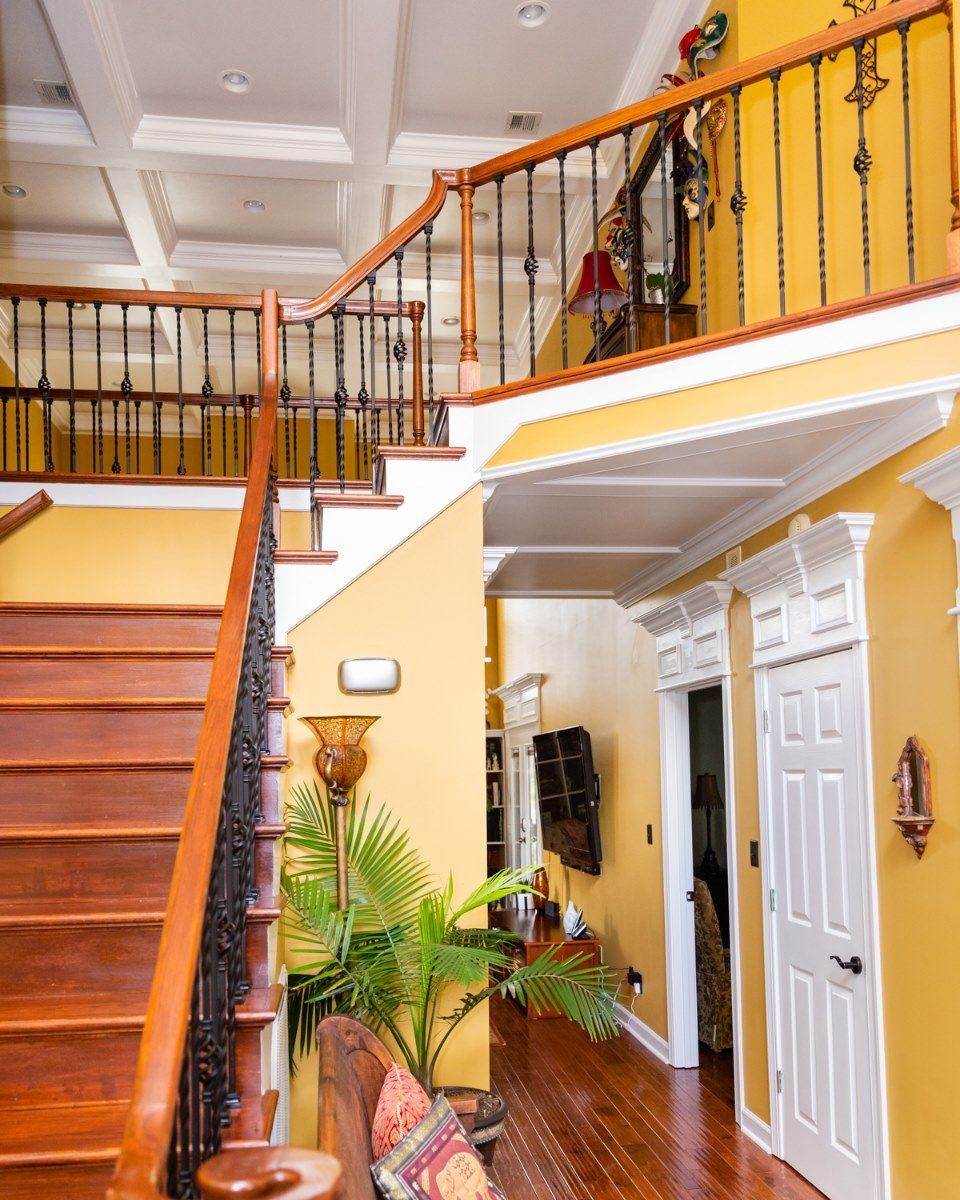 Interior view of a foyer with wooden stairs leading up, yellow walls, a houseplant, and a hallway with two white doors.