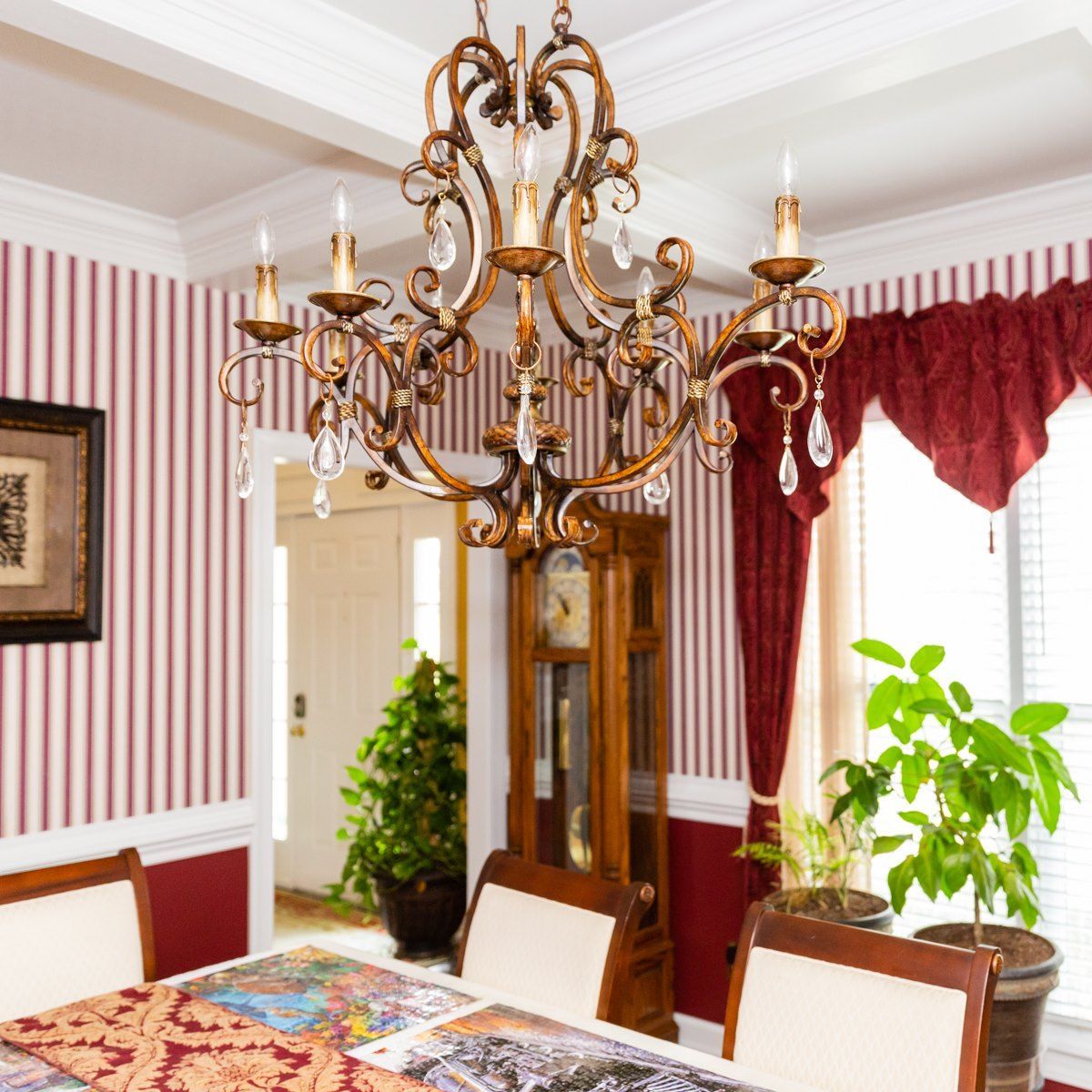 An ornate gold chandelier hangs above a dining table in a room with striped wallpaper and red curtains.