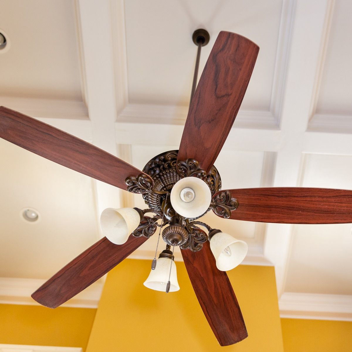 A ceiling fan with five dark wood blades and four glass lights hangs against a white coffered ceiling above yellow walls.