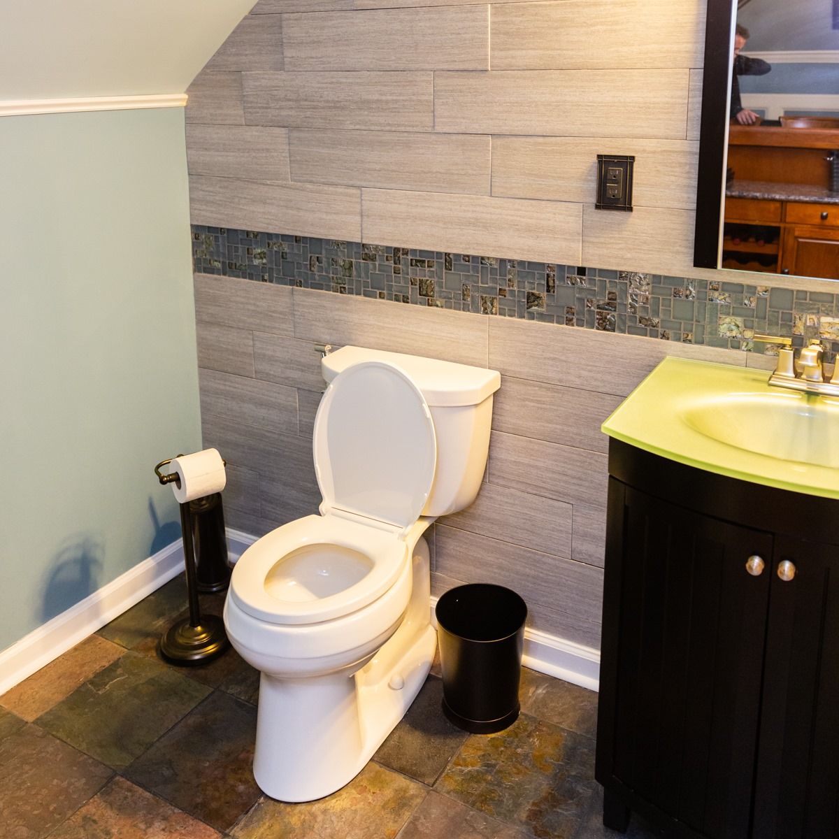 A bathroom featuring a white toilet, slate tile floor, light blue wall, and a gray stone wall with a decorative trim.