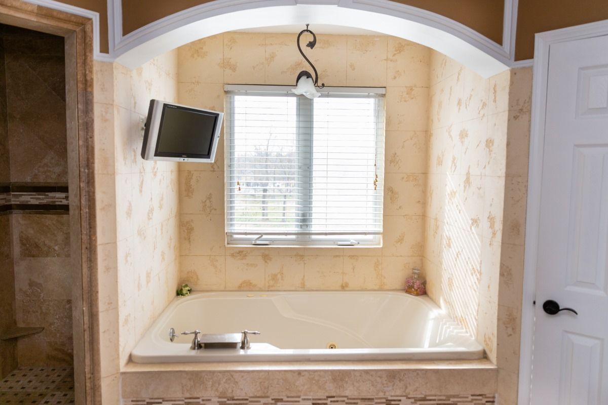 A beige-tiled bathroom alcove featuring a white bathtub, a window with closed blinds, and a small TV on the side wall.