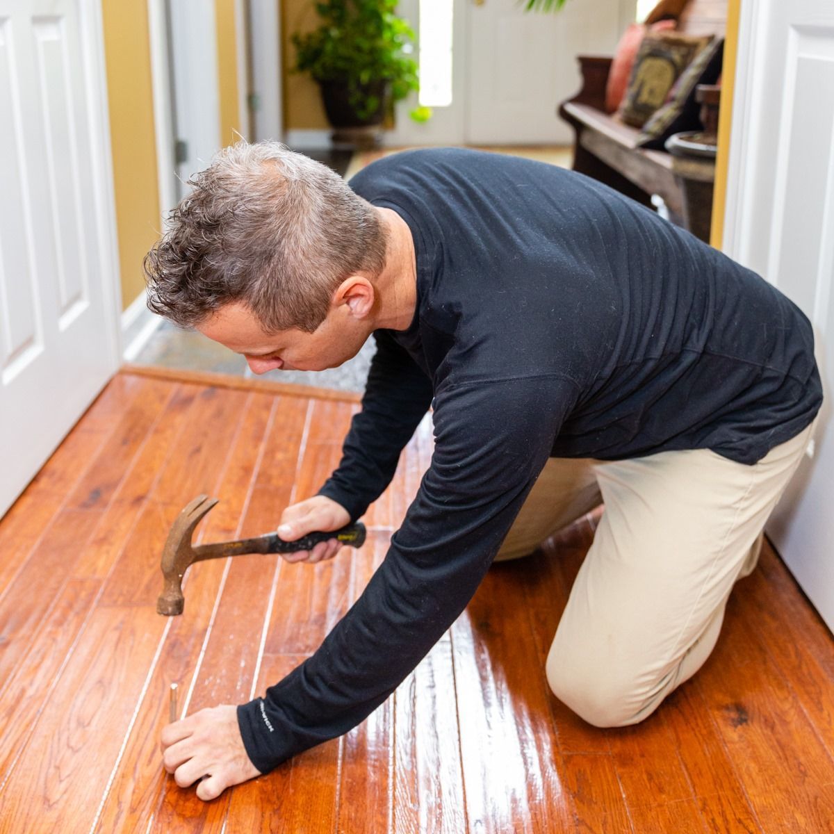 A person kneeling on a wooden floor, using a hammer to secure a small piece of wood between the floorboards.