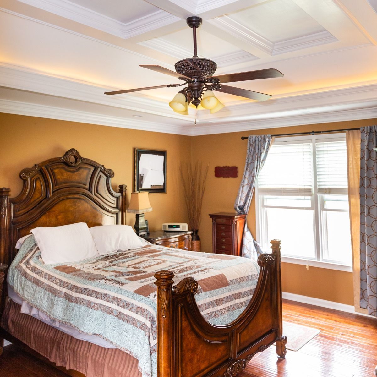 A traditional bedroom with a carved wooden bed frame, patterned quilt, warm-toned walls, and a ceiling fan.