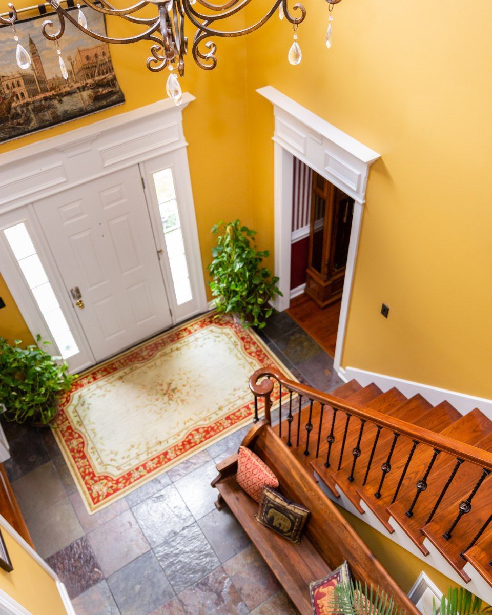 A high-angle view of a sunny foyer with yellow walls, a stone floor, a chandelier, a front door, and a wooden staircase.