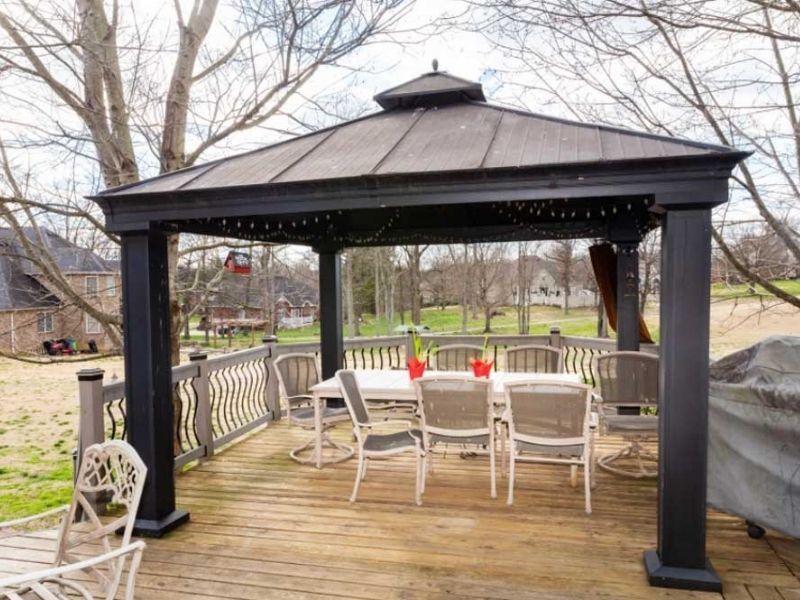 A black outdoor gazebo on a wooden deck featuring a dining table with chairs, set in a backyard during late autumn.