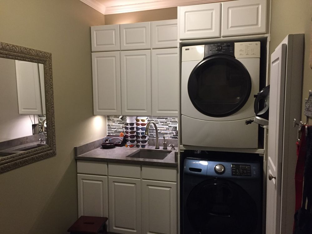 Laundry room with white cabinets, a sink with a mosaic backsplash, a stackable washer and dryer, and a wall mirror.