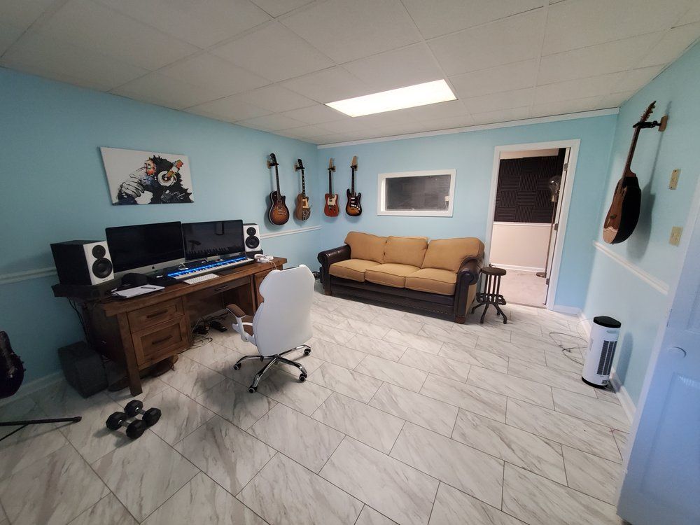 A light blue home studio with a wooden desk, computer setup, a tan sofa, several hanging guitars, and white tile floors.