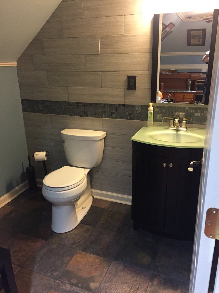 A half bathroom with grey wood-look wall tiles, a mosaic accent strip, dark vanity with glass sink, and a white toilet.