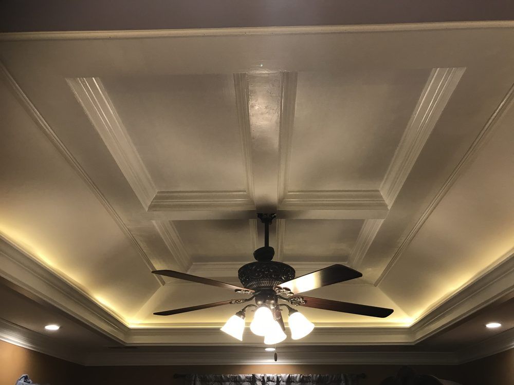 A ceiling fan with a three-light fixture centered on a recessed, coffered ceiling with perimeter ambient cove lighting.