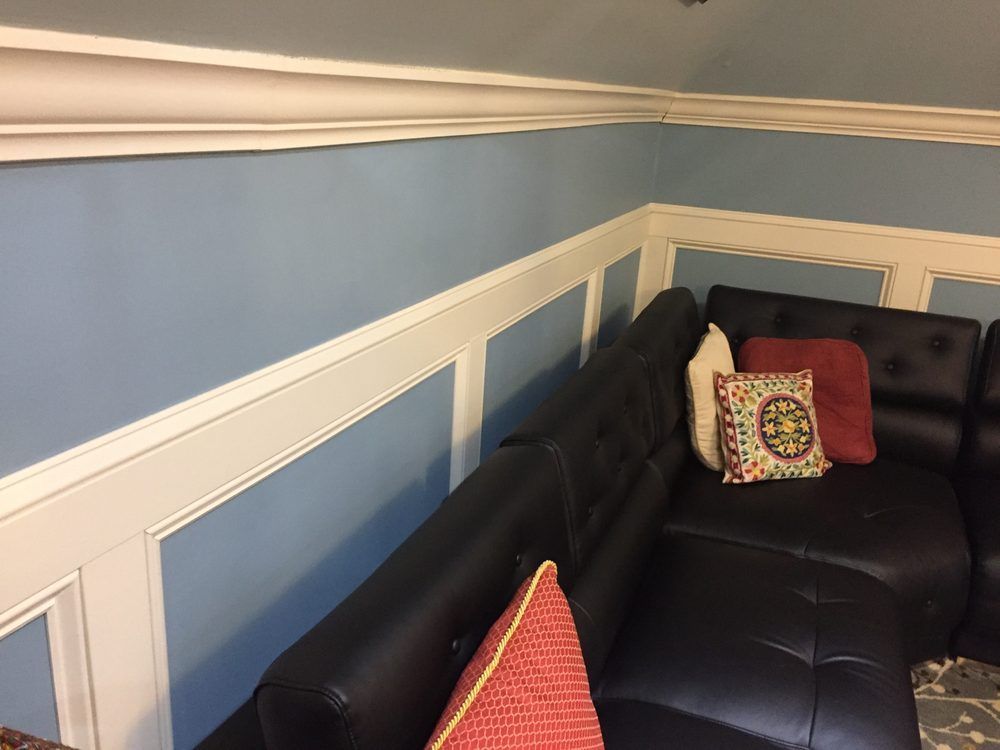 A black leather sectional sofa sits against light blue walls featuring white wainscoting and crown molding.