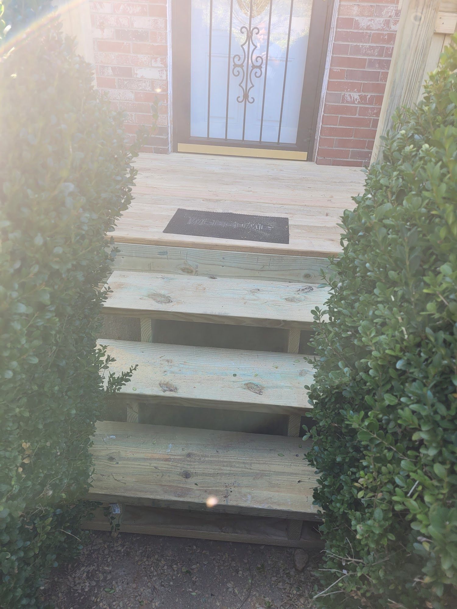 Wooden outdoor steps leading to a brick home's entryway, framed by two large green bushes.