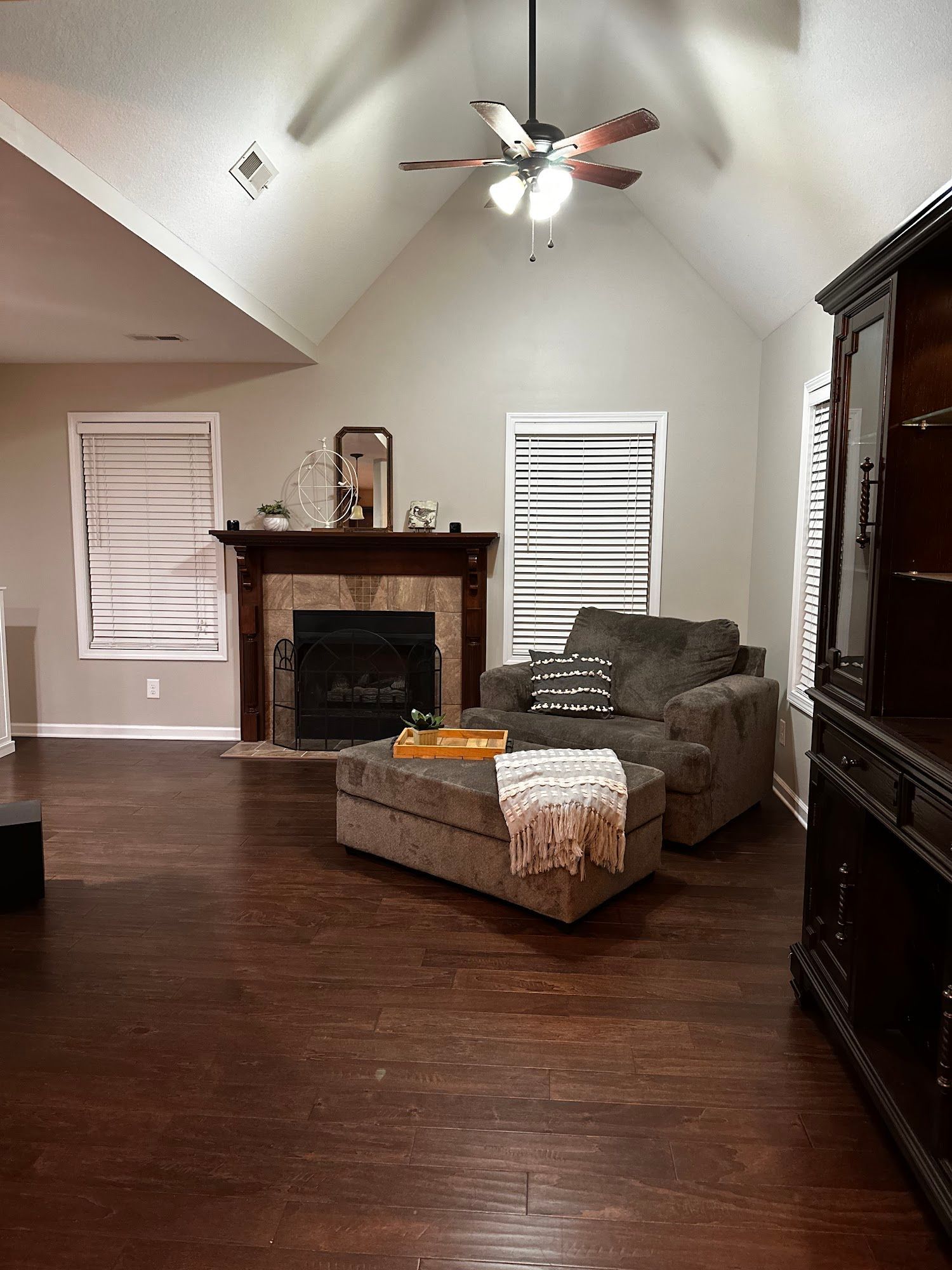 A living room with a stone fireplace, dark wood floors, a ceiling fan, windows with white blinds, and an armchair.