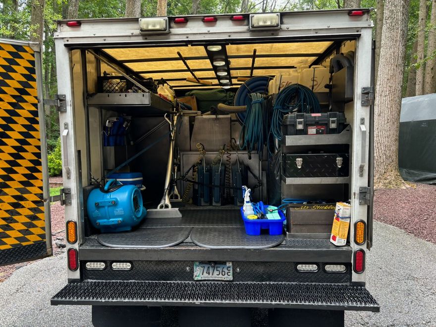 Open utility truck with hoses, tools, and a blue pressure washer in the back, parked outdoors.