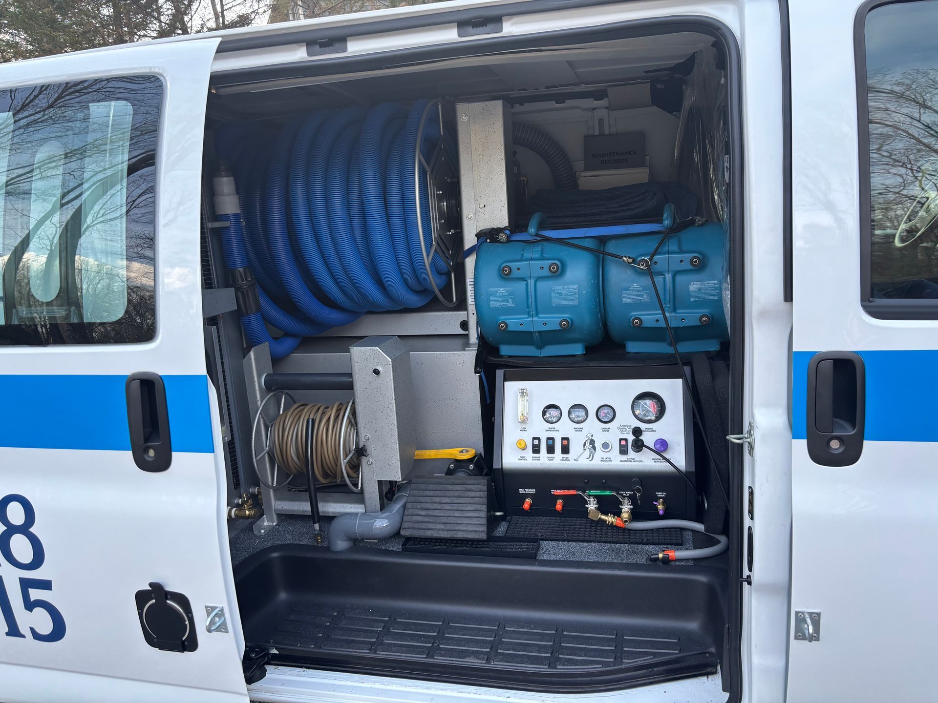 The side door of a white utility van is open, revealing a carpet cleaning machine with blue hoses and control gauges. Open van with water or cleaning equipment, blue hoses and control panel inside