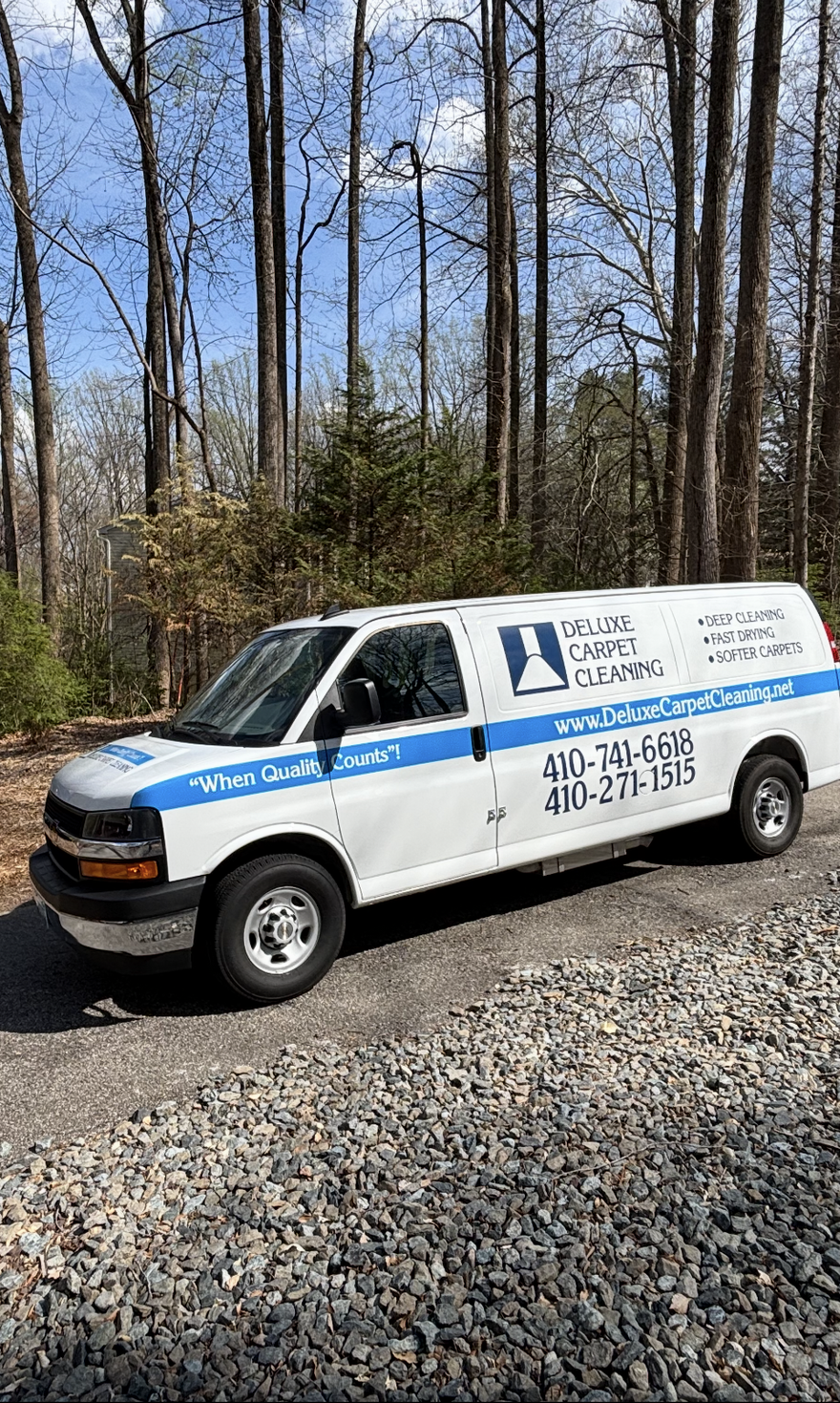 White and blue utility truck parked on gravel in a wooded area, with company branding on the side.