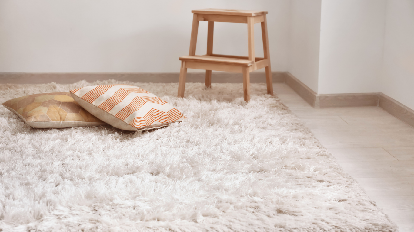 A room with a white rug , pillows and a wooden stool.