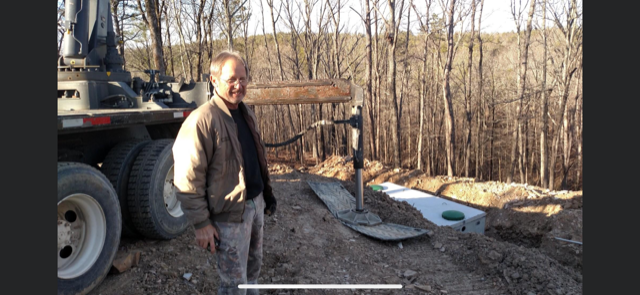 Worker standing beside an excavator in a muddy yard near a wooded area and trench