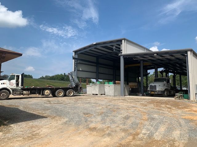 Gravel yard with open metal sheds, trucks, and a flatbed trailer under a blue sky