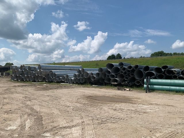 Pipes and large plastic drainage tubes stacked on a dirt lot under a cloudy blue sky