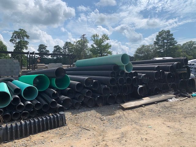 Stacks of black and green plastic pipes stored outdoors on a dirt lot under a cloudy sky