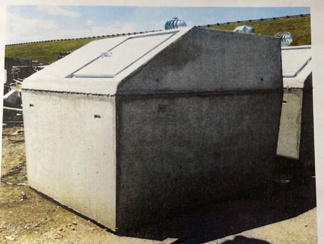 Large gray concrete utility enclosure with a sloped lid outdoors on a grassy hillside.
