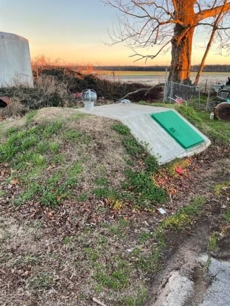Outdoor utility cover beside a grassy mound at sunset, with a green access hatch and nearby tree branches